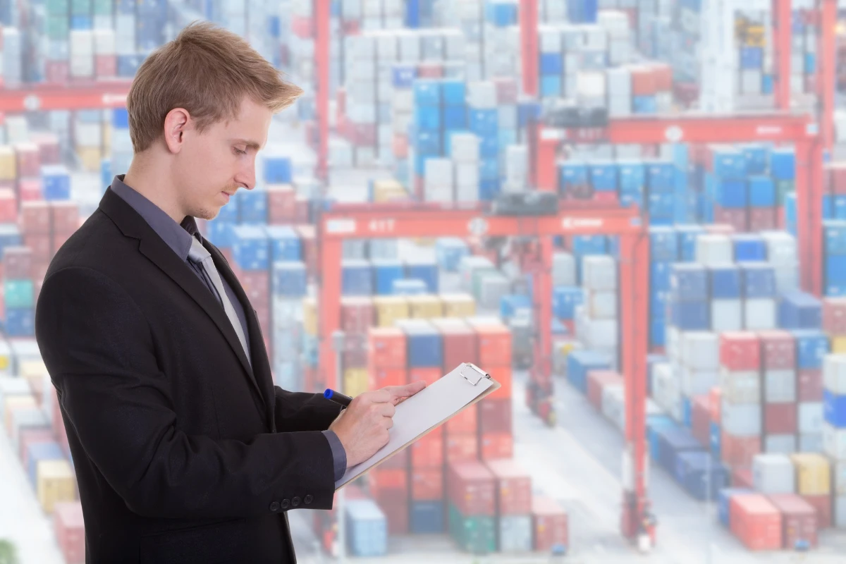 A man in a suit holds a clipboard in front of a container ship at a storage container storage yard.